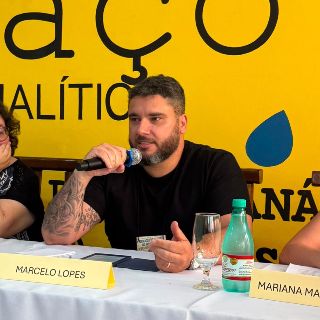 Homem com tatuagens falando em um microfone durante um painel; ao fundo, uma parede amarela com texto. À esquerda, mulher ouvindo atentamente.